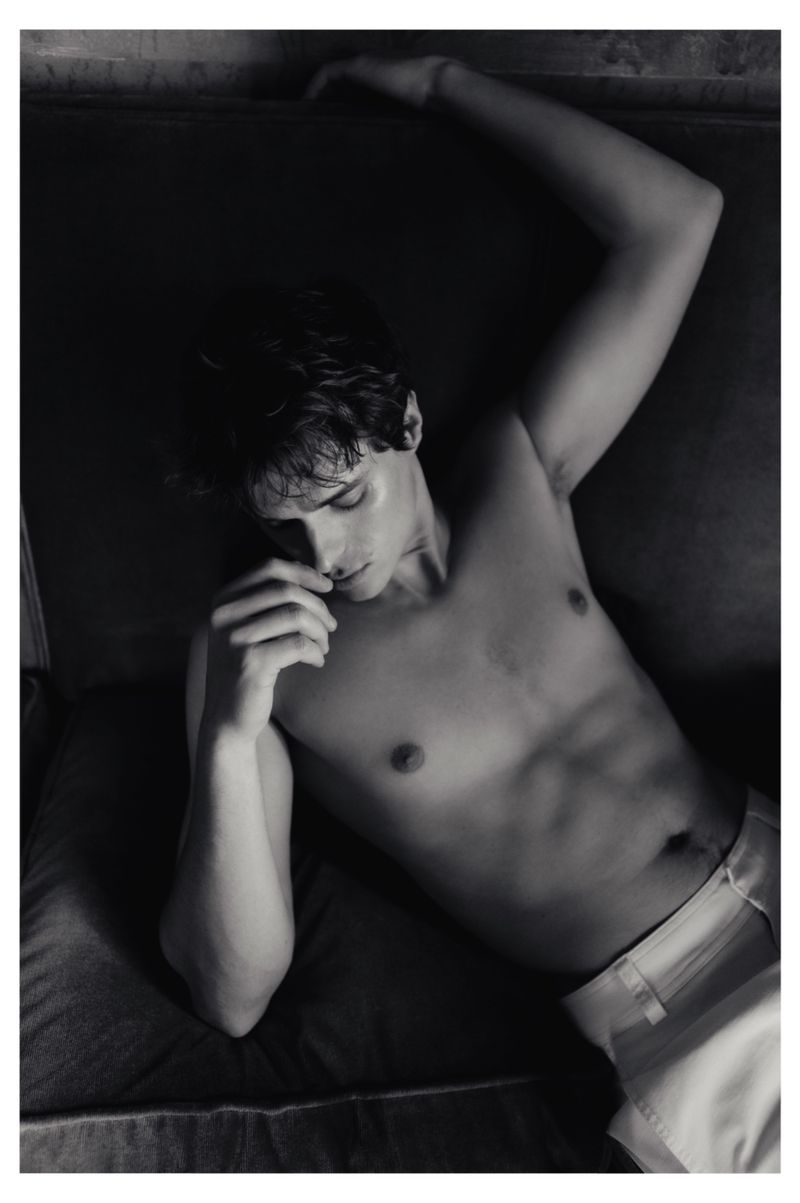 Shirtless young man reclining indoors wearing light trousers in a monochrome editorial portrait.