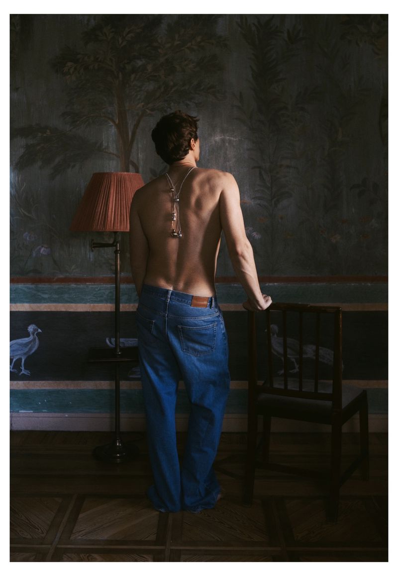 Shirtless man standing with his back turned, wearing blue denim jeans and a long silver necklace draped down his spine.