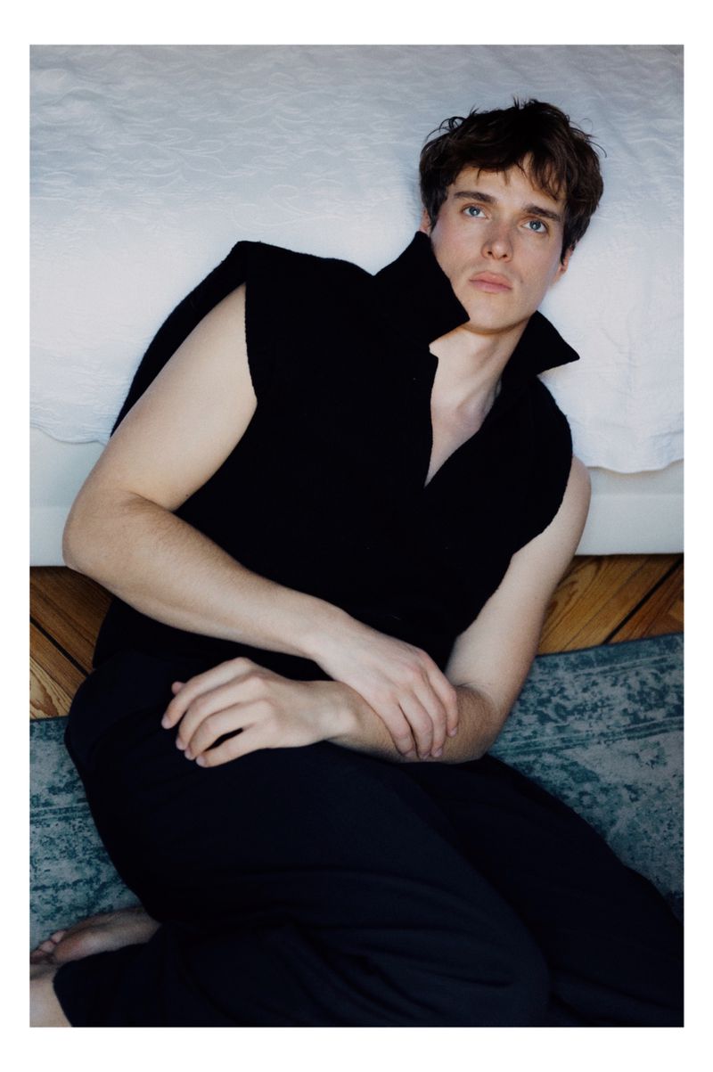 A young adult man is lying on a light-colored bed with a green rug on the floor in a medium shot, wearing a black outfit.