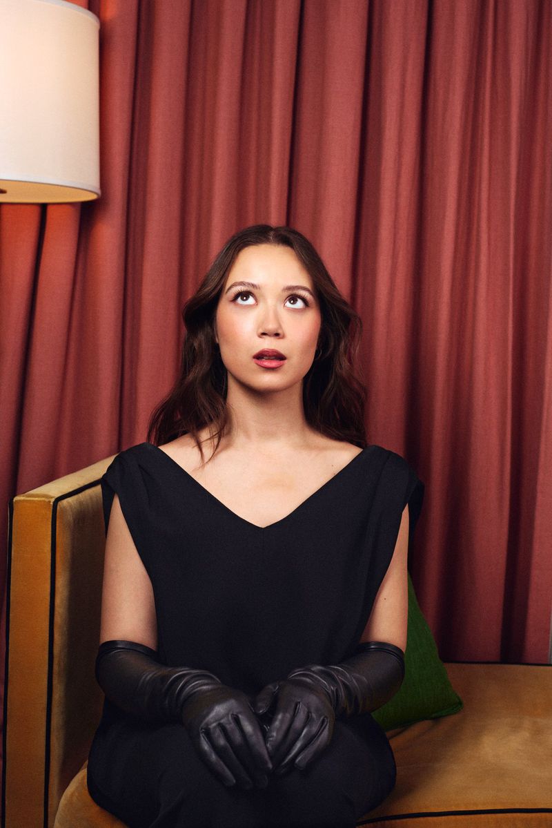 Portrait of Laufey in a black top and leather gloves looking upward, seated on a mustard velvet sofa against draped red curtains.