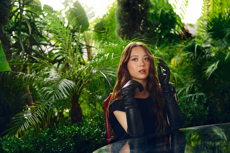 Laufey seated amidst lush tropical foliage wearing black leather gloves for Billboard Magazine