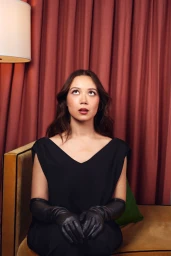 Portrait of Laufey in a black top and leather gloves looking upward, seated on a mustard velvet sofa against draped red curtains.