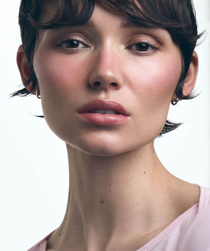 Close-up portrait of a woman with short dark hair and natural makeup wearing a light pink top against a plain white background.