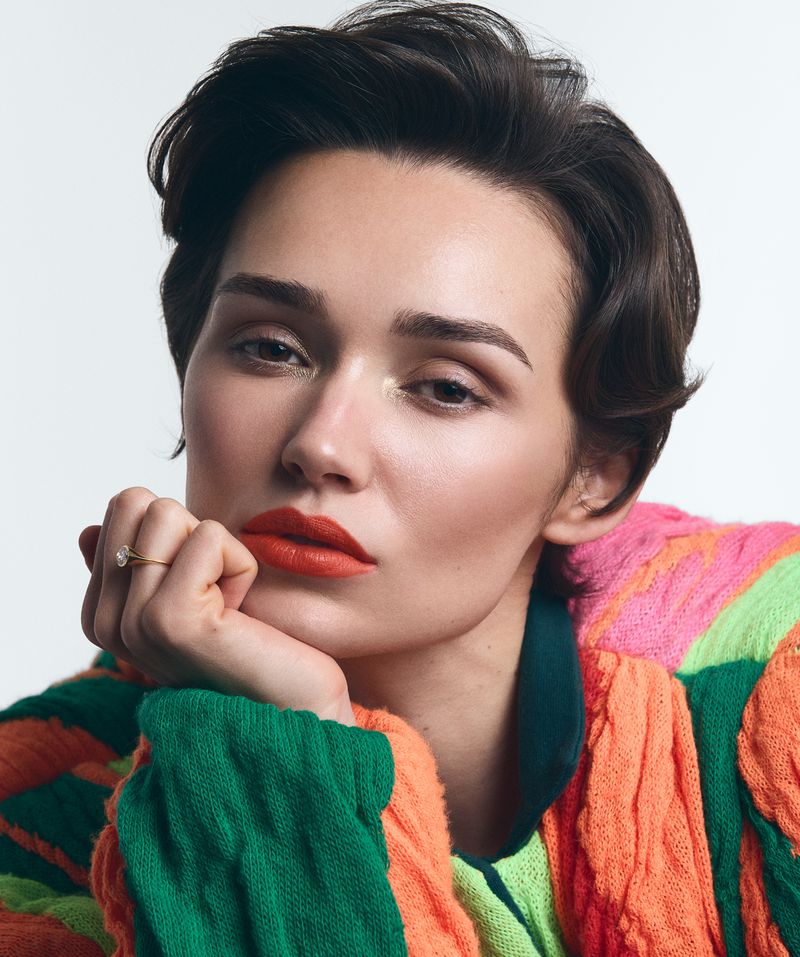 Close-up portrait of a young adult woman with short dark hair, light skin, and red lips, wearing a bright colorful knit sweater.