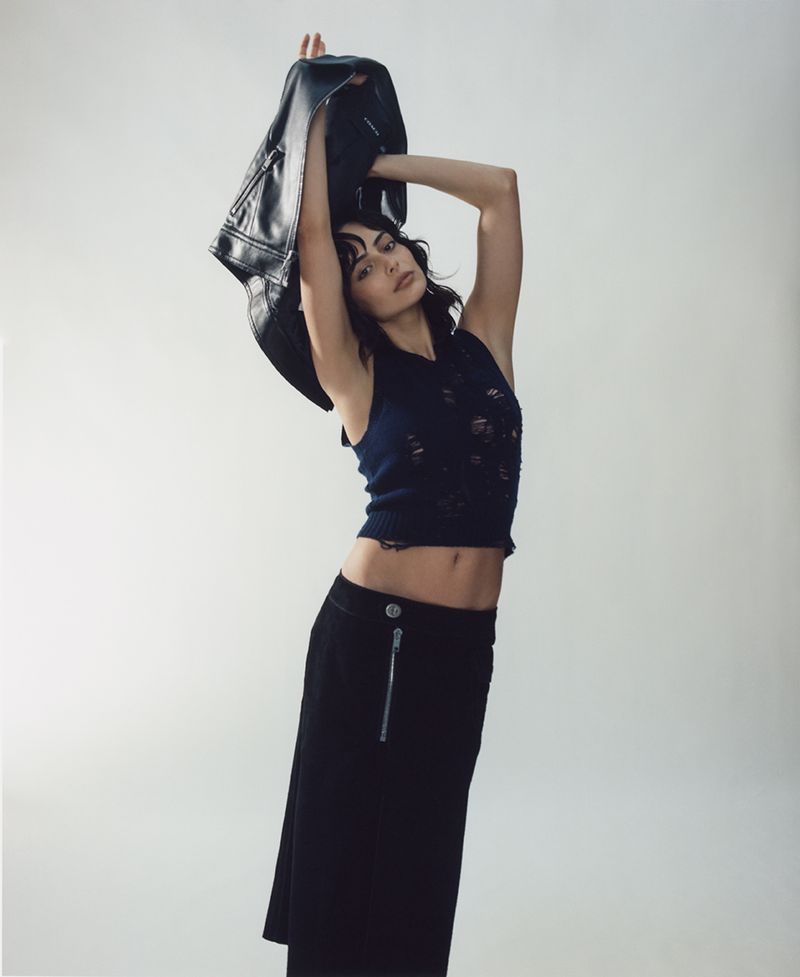 Indoor fashion portrait of adult female wearing a dark-colored tank top and midi skirt, holding a leather jacket overhead against a grey backdrop.