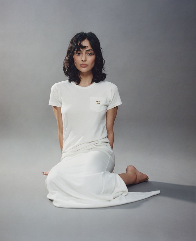 Woman with dark hair wearing a white short-sleeve t-shirt and white skirt seated on a grey seamless background