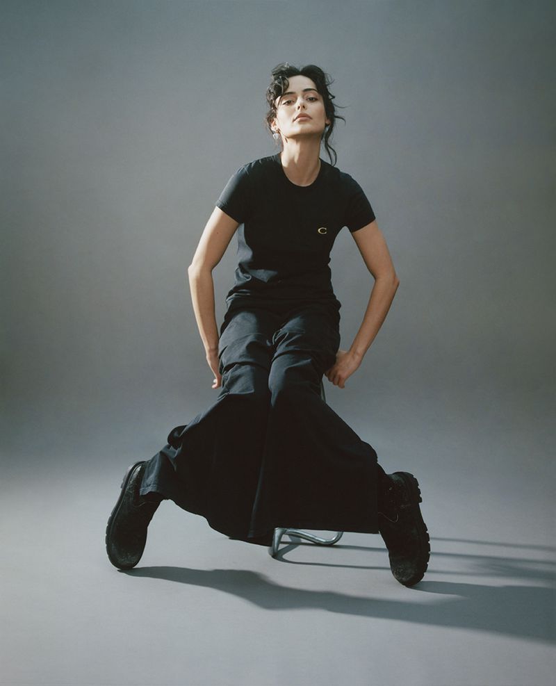 Woman wearing a black t-shirt and black trousers seated on a stool against a grey studio backdrop.