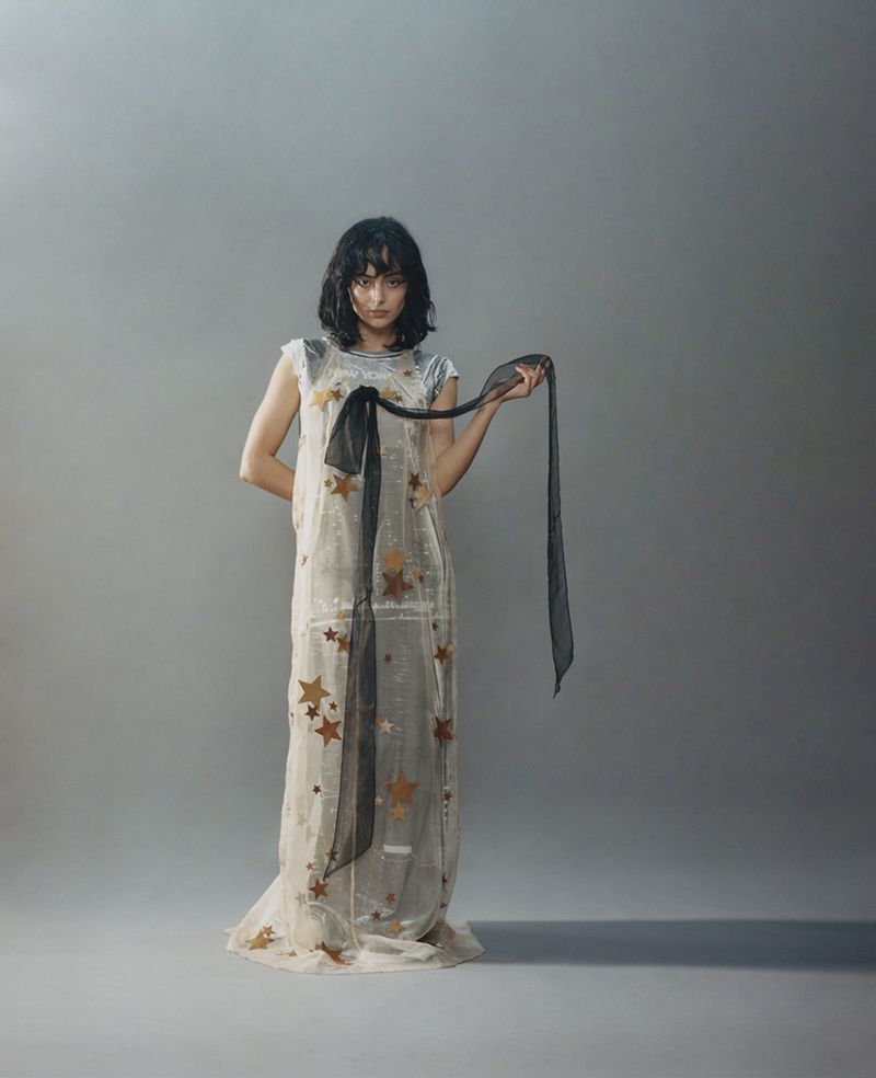 Woman in a sheer star-patterned dress holding a black ribbon against a grey seamless background
