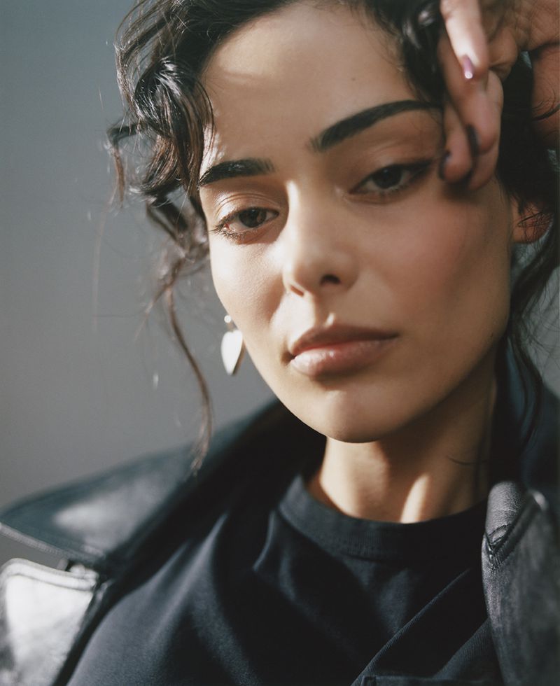 Close-up portrait of a woman with dark wavy hair wearing a black t-shirt and leather jacket against a grey background