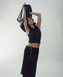 Woman in dark sleeveless top holding a black leather jacket overhead against a neutral seamless background.