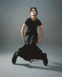 Woman wearing a black t-shirt and black trousers seated on a stool against a grey studio backdrop.