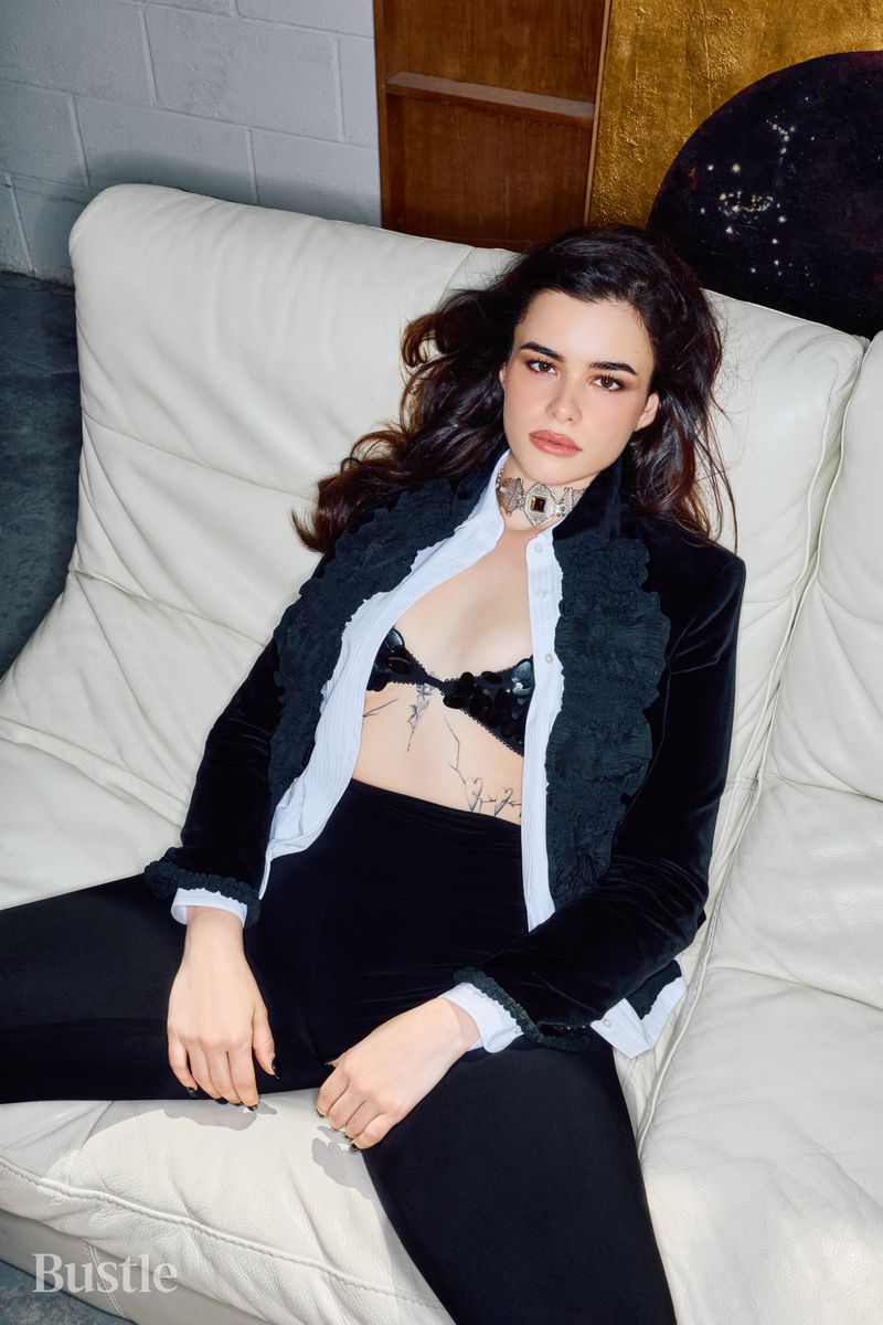 Barbie Ferreira lounging on a white leather sofa wearing a black velvet jacket over a white shirt and black trousers.