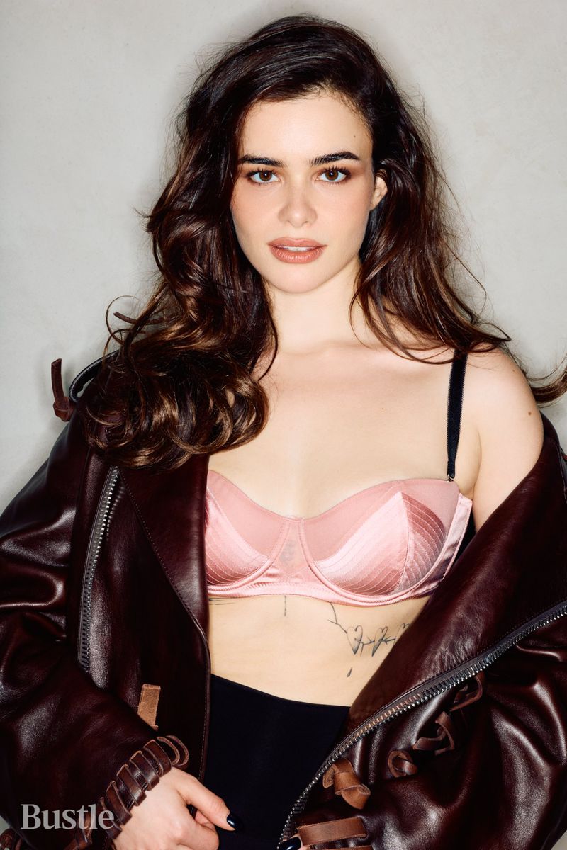 Portrait of Barbie Ferreira wearing a pink bra and brown leather jacket against a neutral grey wall