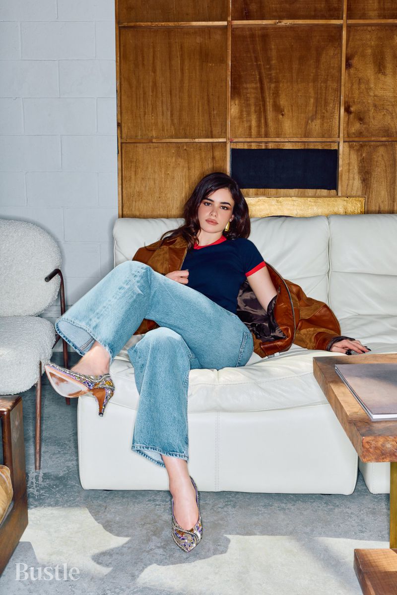 A woman with dark hair sits on a white couch wearing jeans, a navy t-shirt, brown jacket, and patterned heels in a wood-paneled room.