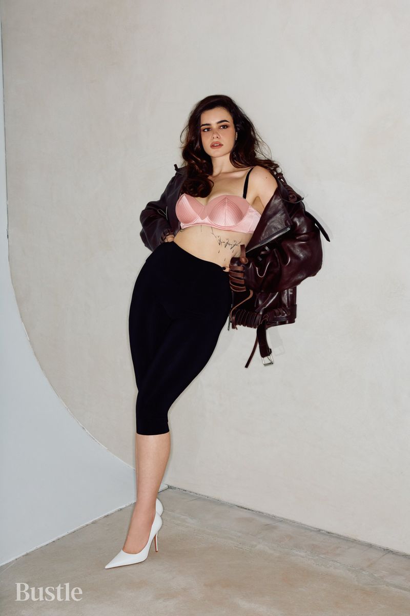 Barbie Ferreira posing against a curved studio wall wearing a pink satin top and dark leather jacket