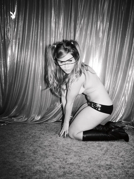 Model Lexee Smith kneeling in black briefs and knee-high boots against a shiny curtain backdrop