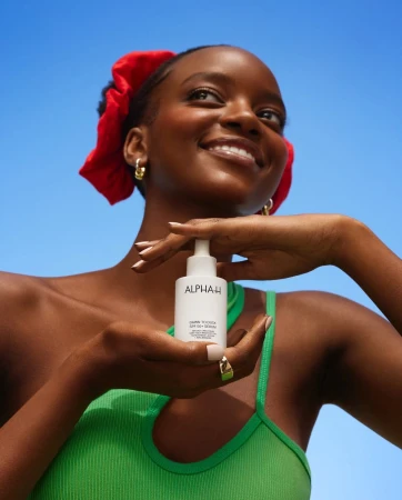 Dark-skinned woman smiling outdoors with a red scrunchie holds Alpha-H Vitamin C serum against a blue sky in a beauty product shot.