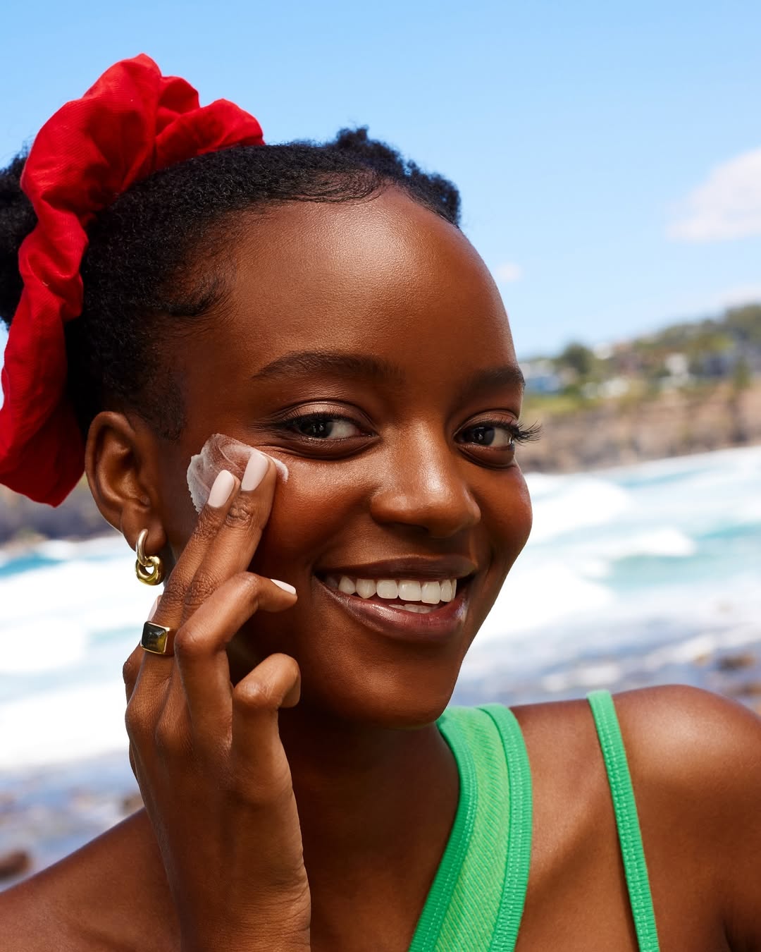 A smiling African-American woman applies sunblock to her face in an outdoor setting with ocean and greenery in the background on a sunny day.