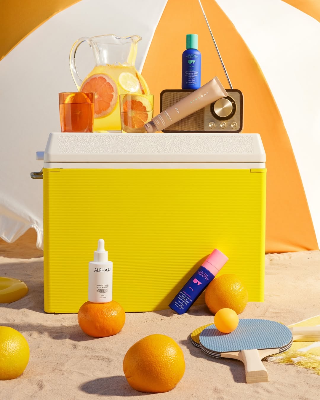 Still life shot of skincare products, citrus fruit, beverage pitcher and ping pong equipment arranged on a sandy surface.