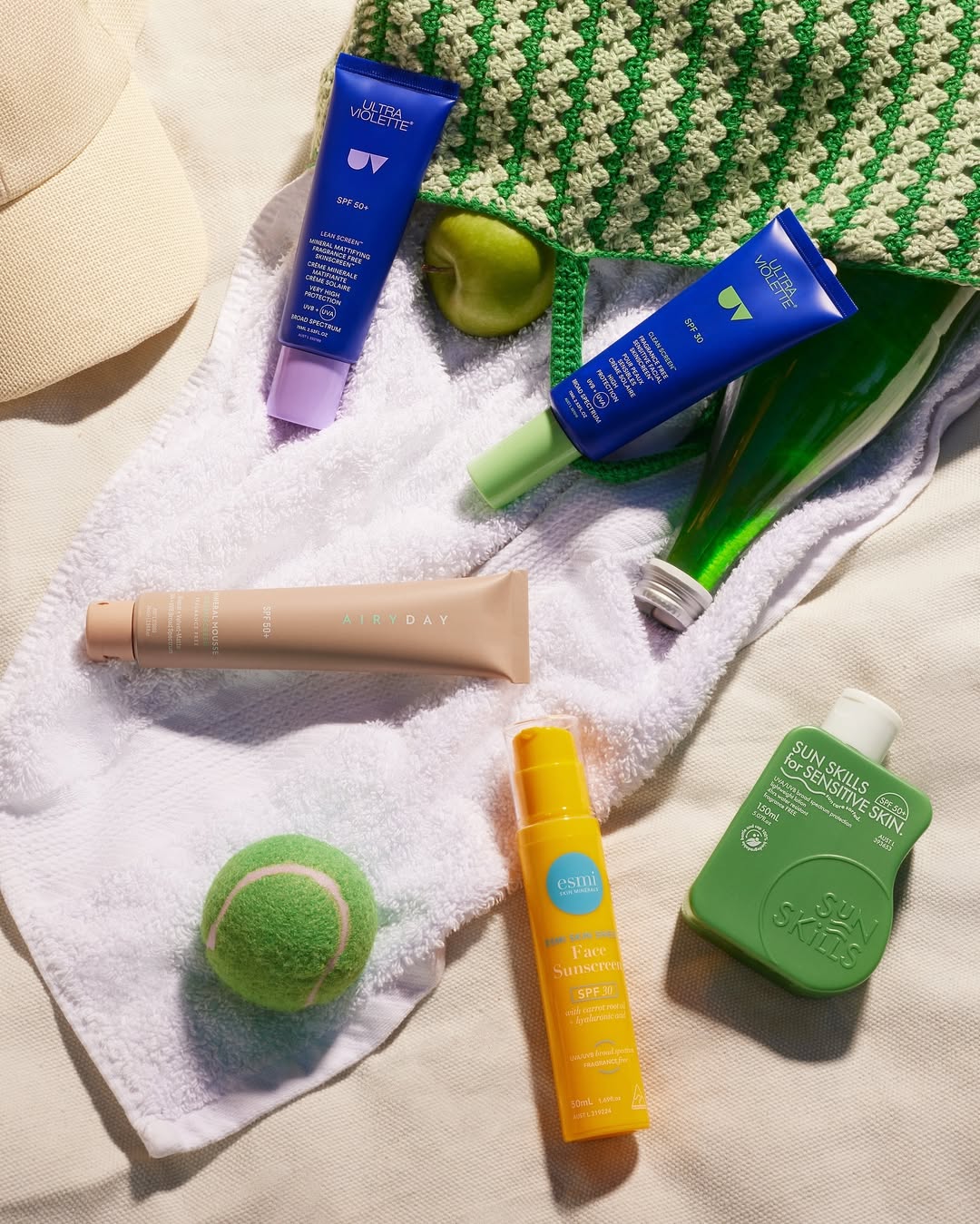Assorted sunscreen bottles and tubes on a white towel next to a green tennis ball, with a tan baseball cap and a crocheted tote bag.