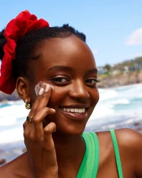 Close-up of a black woman applying white sunscreen to her cheek with a red scrunchie in her hair and a green top by the ocean