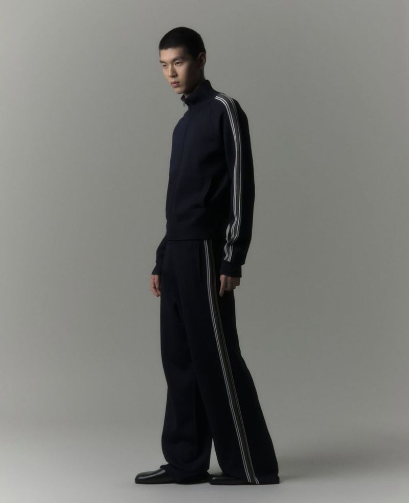 Studio shot of an Asian man in a dark tracksuit with white stripes, standing in a simple pose against a solid grey backdrop.
