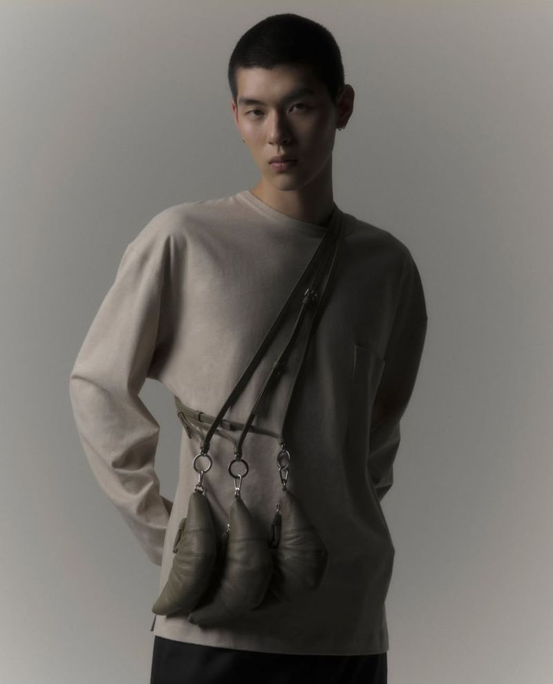 Asian man poses in front of a neutral background wearing a beige long-sleeved shirt with a unique bag that has pod-like attachments.
