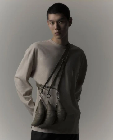 Asian man poses in front of a neutral background wearing a beige long-sleeved shirt with a unique bag that has pod-like attachments.