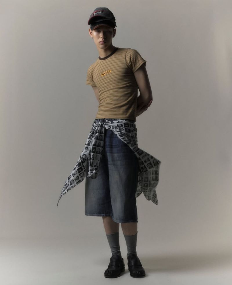 A slender young adult poses indoors, wearing a striped tan t-shirt, denim shorts, and a baseball cap, styled with neutral gray backdrop.