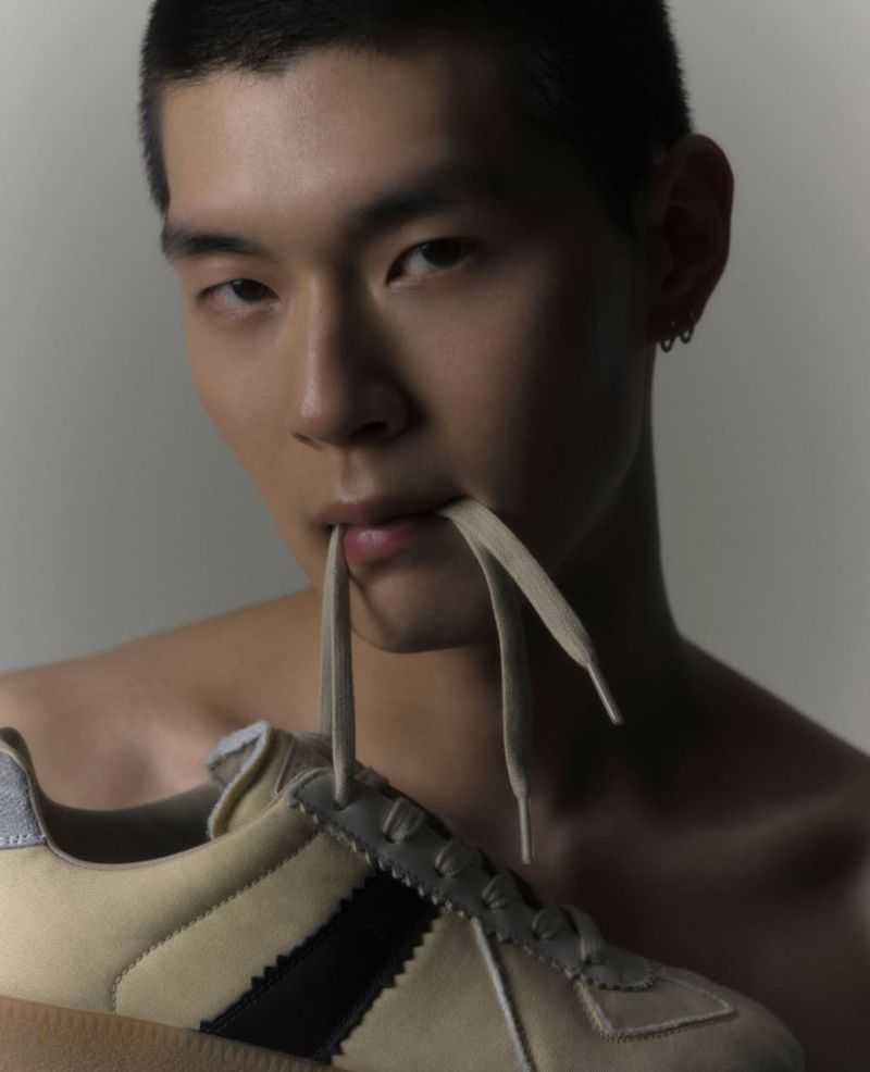 Portrait of a shirtless Asian man holding a sneaker in front of him with the shoelaces in his mouth against a neutral background.