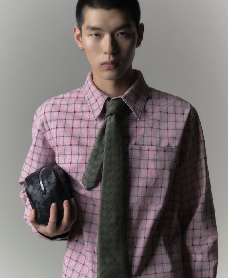 Young man in a pink shirt and green tie, holding a black woven clutch, in a studio portrait with soft lighting and neutral background.