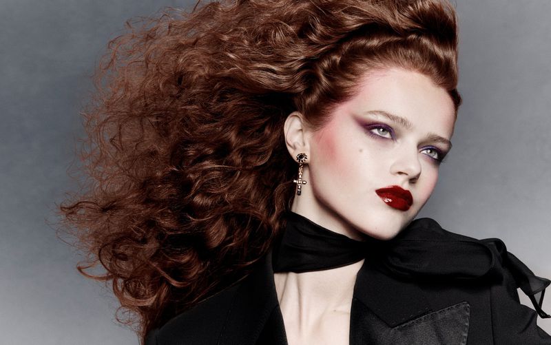 Model Charlotte Boggia with voluminous red hair wearing dark lipstick and a black blazer against a grey studio backdrop.