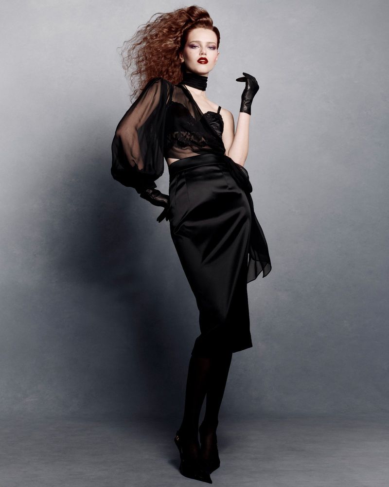 Model Charlotte Boggia posing in a black sheer top and satin skirt against a grey seamless studio backdrop.