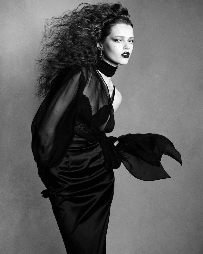 Model Charlotte Boggia with voluminous dark hair wearing a black sheer blouse against a neutral studio backdrop.