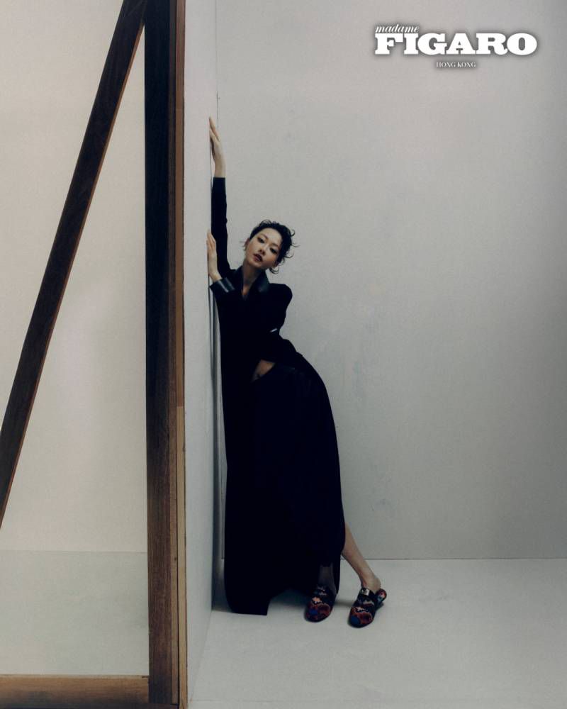 Woman in black outfit leaning against a white studio wall with wooden architectural frames