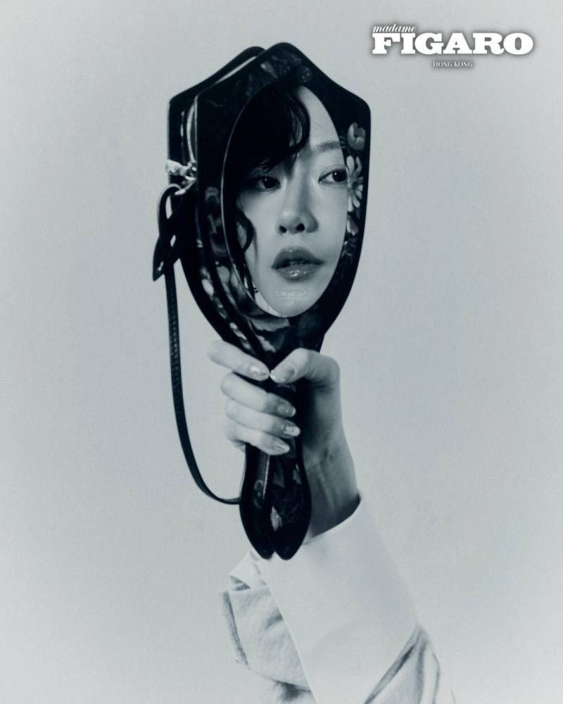 Monochrome editorial portrait of a woman reflected in a hand mirror held up against a plain white background.