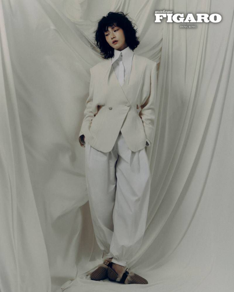 Woman with short dark hair wearing an off-white blazer and matching trousers against a draped fabric backdrop.