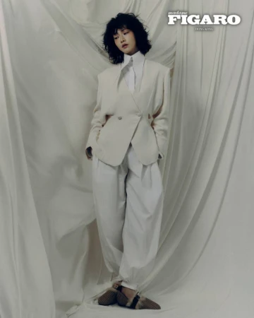 Woman with short dark hair wearing an off-white blazer and matching trousers against a draped fabric backdrop.