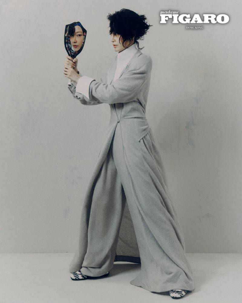 Woman in a long grey coat holding a small ornate hand mirror against a plain light grey backdrop