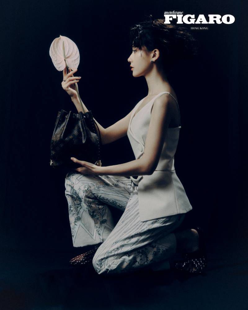 Fish Liew kneeling in profile wearing a white top and patterned trousers holding a white flower and dark handbag against a dark background