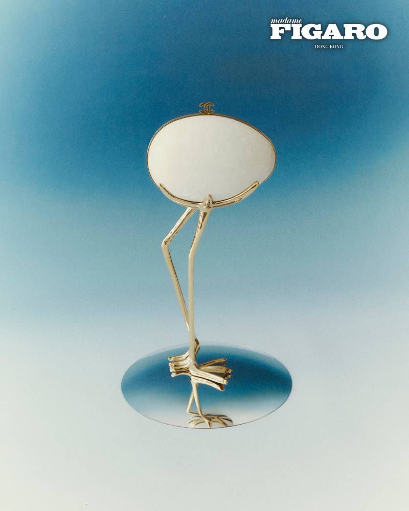 An ovoid white handbag with slender golden bird legs standing on a reflective circular base against a blue gradient background.