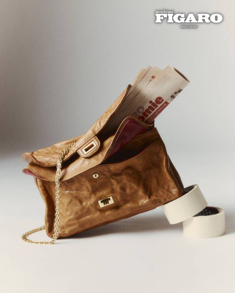 Tan leather handbag with folded newspapers protruding from its open flap against a light background