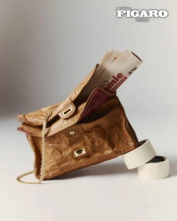 Tan leather handbag with folded newspapers protruding from its open flap against a light background
