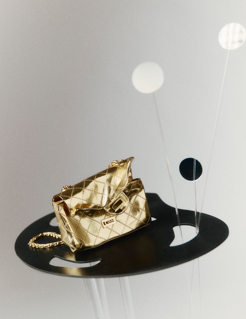 Gold quilted handbag with chain strap resting on an abstract black platform with wire floral sculptures against a pale grey background