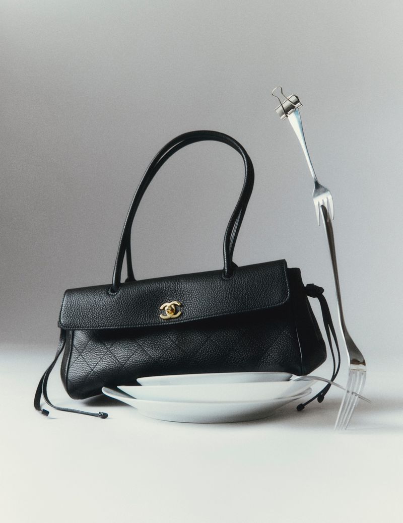 Black leather handbag resting on white plates with a bent metal fork secured by a binder clip against a pale grey background.