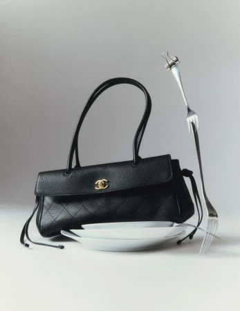 Black leather handbag resting on white plates with a bent metal fork secured by a binder clip against a pale grey background.