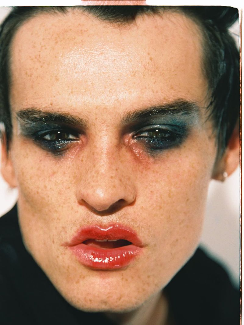 Close-up portrait of a male model with heavy dark eye makeup and glossy red lips against a neutral background.