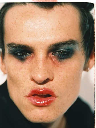 Close-up portrait of a male model with heavy dark eye makeup and glossy red lips against a neutral background.