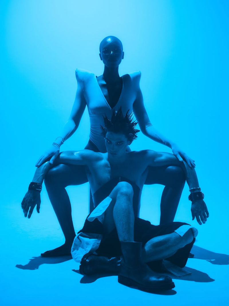 Two models posed against a vibrant blue studio backdrop with dramatic monochromatic lighting and avant-garde styling.