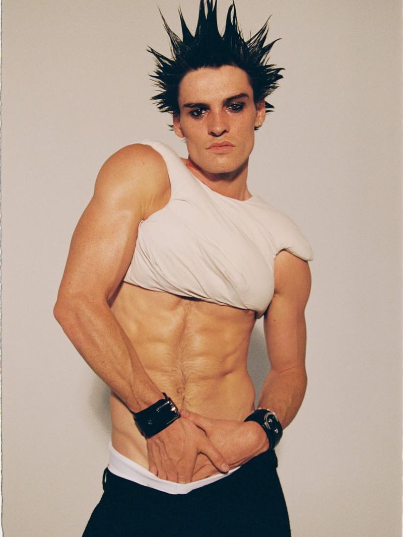 A male model with spiked dark hair and heavy eyeliner wearing a white crop top and black trousers against a neutral background